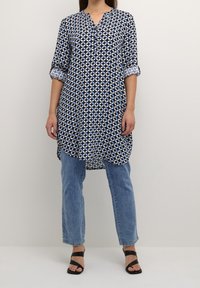 Tunic navy con motivo geometrico bianco e blu, scollatura a V, maniche arrotolate a 3/4, abbinata a jeans chiari e sandali neri con cinturini.