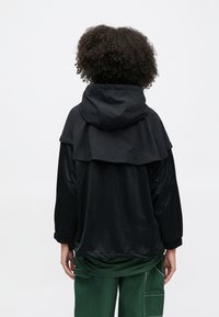 Μαύρο μπουφάν με κουκούλα και στρωματοποιημένο σχέδιο, ελαστικά μανίκια και γυαλιστερή υφή. Φορεμένο πάνω από πράσινο παντελόνι cargo.