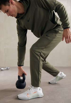 Mand i grønt træningstøj løfter en sort kettlebell, iført hvide sneakers med orange Nike-logoer på en grå gulv.