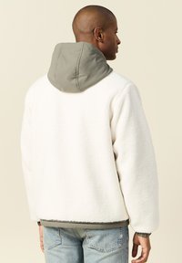 Biała polarowa kurtka z szarym kapturem, charakteryzująca się luźnym krojem i ściągaczami, prezentowana od tyłu z gładką fakturą.