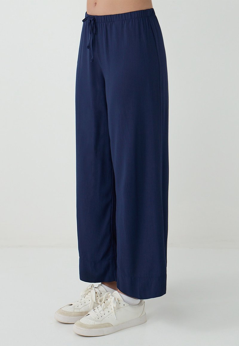 Marineblauwe wijde broek met een elastische tailleband en trekkoord. De stof heeft een gladde textuur. Draagt bij witte sneakers en enkelkousen.