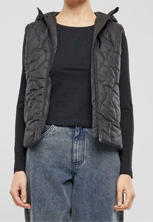 Persoon draagt een zwart doorgestikt mouwloos vest over een zwarte top met lange mouwen en een hoog uitgesneden vervaagde blauwe spijkerbroek, staand met ontspannen armen.