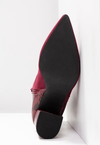 Chaussure rouge à bout pointu avec motif texturé et semelle noire reposant verticalement contre un coin blanc du mur et du sol.