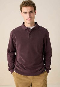 Mann mit lockigem Haar, der ein dunkelviolettes langärmeliges Polohemd und beige Cordhosen trägt, steht mit den Händen in den Taschen vor einem neutralen Hintergrund.