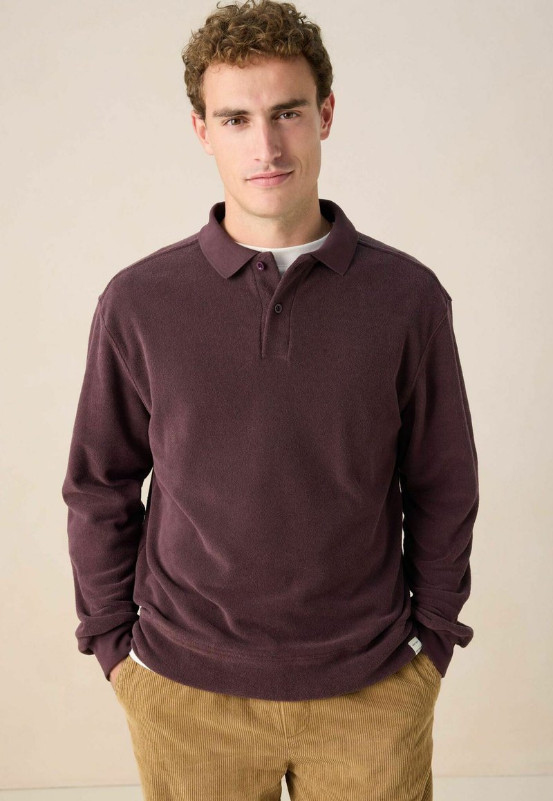 Mann mit lockigem Haar, der ein dunkelviolettes langärmeliges Polohemd und beige Cordhosen trägt, steht mit den Händen in den Taschen vor einem neutralen Hintergrund.