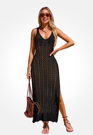 Woman wearing black sleeveless crochet maxi dress, brown braided handbag, brown sandals, sunglasses, and gold jewelry, smiling with hand on hip.