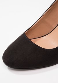 Chaussure à talon haut en suède noir avec un bout pointu, présentant une texture lisse et un intérieur en nude. Design simple et élégant.