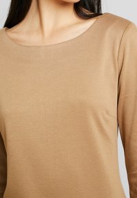 Maglia a maniche lunghe color nocciola chiaro, con una texture morbida e liscia e un collo ampio e arrotondato. Presenta un design minimalista e senza decorazioni visibili.