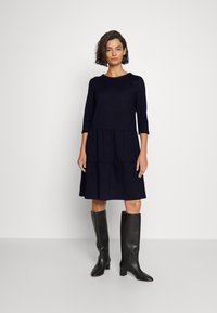 Marineblaues, knielanges Kleid mit Dreiviertelärmeln und gestuftem Design. Getragen mit schwarzen knielangen Stiefeln mit spitzer Zehe und Blockabsatz.