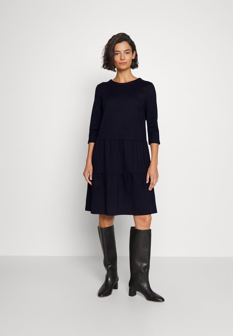 Marineblaues, knielanges Kleid mit Dreiviertelärmeln und gestuftem Design. Getragen mit schwarzen knielangen Stiefeln mit spitzer Zehe und Blockabsatz.