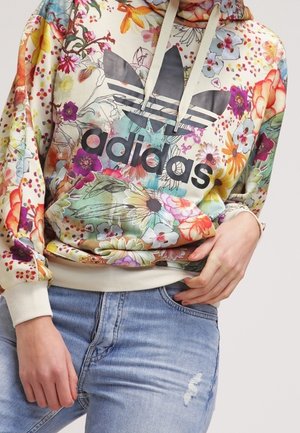 Person iført en farverig blomster Adidas hættetrøje med stort sort logo og lyseblå revnede jeans, hænderne hvilende nær taljen.