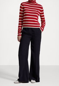 Personne portant un pull à col montant rayé rouge et blanc, un pantalon large noir et des ballerines blanches, debout avec une main dans la poche.