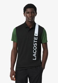 Polo shirt nero e verde a maniche corte, con una striscia verticale bianca e il testo "LACOSTE". Realizzato in tessuto traspirante.