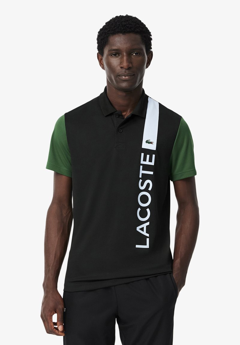 Polo shirt nero e verde a maniche corte, con una striscia verticale bianca e il testo "LACOSTE". Realizzato in tessuto traspirante.