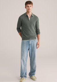 Zelen pulover iz pletenine z ovratnikom, v kombinaciji s svetlo modrimi širokimi jeans hlačami in bež čevlji, stoji na nevtralnem ozadju.