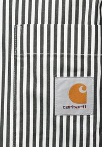 Camisa de rayas en blanco y negro con un bolsillo de parche. Presenta una etiqueta cuadrada con un logo naranja y texto burdeos, confeccionada con un tejido duradero.