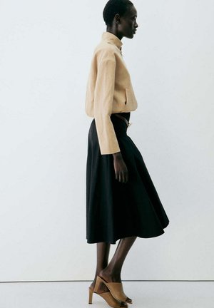 Mujer caminando con un top beige de manga larga, falda midi negra y zuecos abiertos de tacón alto color marrón claro, sobre un fondo blanco liso.