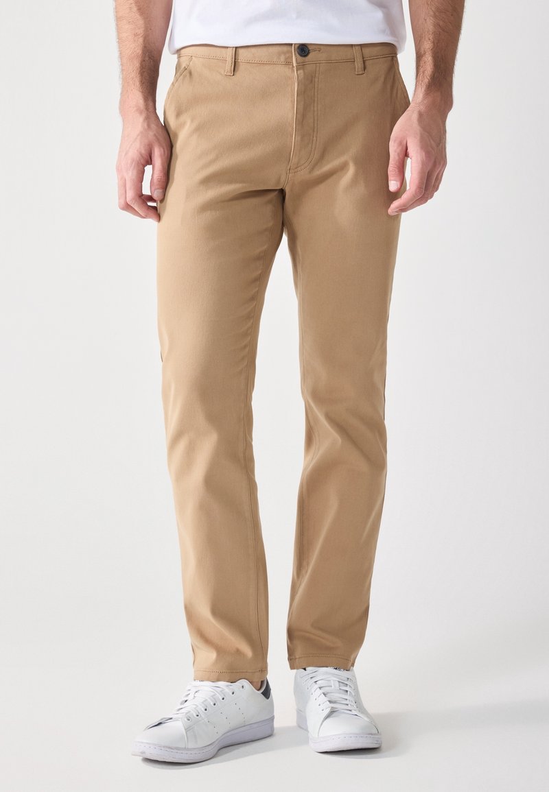 Homme portant un pantalon beige ajusté et des baskets blanches, debout devant un fond uni.