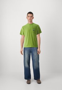 Zeleni bombažni T-shirt z okroglim izrezom in kratkimi rokavi, ki ima prsni žep z temno modrim oznako. Obučen z modrimi širokimi hlačami.