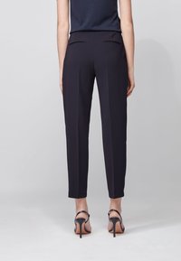 Černé přiléhavé kalhoty vyrobené z hladké tkaniny, se zúženými nohavicemi, jemnými zadními kapsami a středně vysokým pasem. Nosené s lodičkami na průhledných podpěrách.