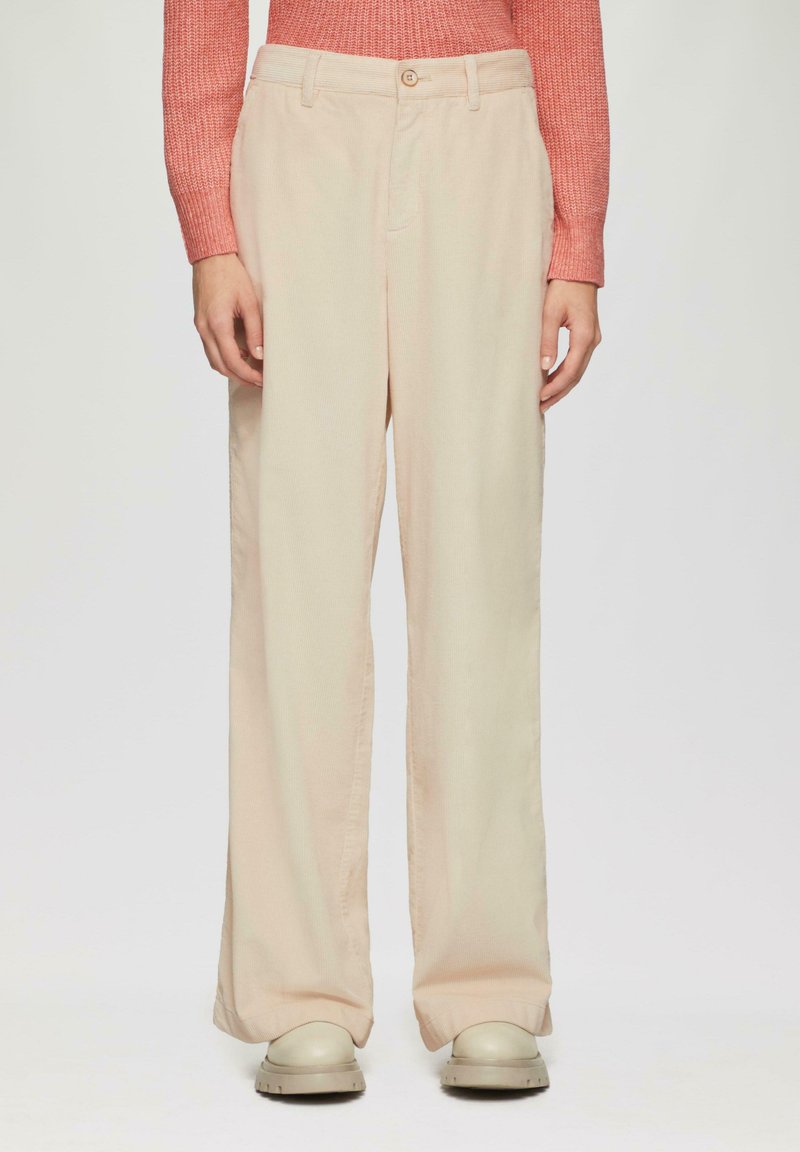 Persoon in een wijde beige broek, witte chunky schoenen en een lange mouw gestreepte koraalkleurige trui, staande tegen een effen achtergrond.