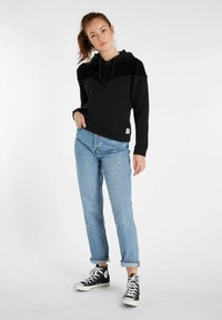 Zwarte hoodie met een corduroy bovenkant, gecombineerd met lichtblauwe opgerolde jeans en zwarte hoge sneakers.