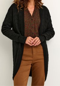 En kvinna bär en svart cardigan med öppna framsidor, en brun mönstrad knäppskjorta och bruna byxor, stående mot en enkel ljus bakgrund.