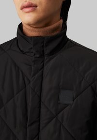 Chaqueta acolchada negra con cuello alto, que presenta una superficie texturizada y un parche cuadrado del logo de Calvin Klein en el pecho.