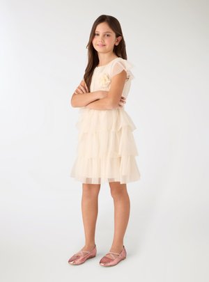 Ragazza in piedi con le braccia incrociate, vestita con un abito crema a più strati e ballerine rosa glitterate su uno sfondo bianco semplice.