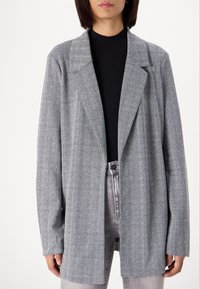 Gray herringbone blazer with a relaxed fit, notched lapels, and long sleeves. Worn over a black top, features a smooth texture and minimal design.