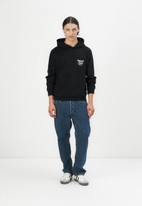 Black cotton hoodie with a front pocket, featuring white text detailing. Paired with loose-fit blue denim jeans and white sneakers.