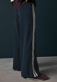 Pantalons larges de couleur marine en tissu doux, avec des rayures blanches et bleu clair sur les côtés. Associés à des baskets bourgogne.