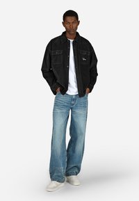 Czarna kurtka dżinsowa z dwoma kieszeniami na piersiach na białej koszulce, zestawiona z jasnoniebieskimi szerokimi dżinsami i białymi trampkami.