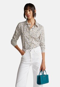 Donna con capelli scuri e lisci, che indossa una camicia a bottoni floreale e pantaloni bianchi, mentre tiene una piccola borsa verde acqua su uno sfondo semplice.