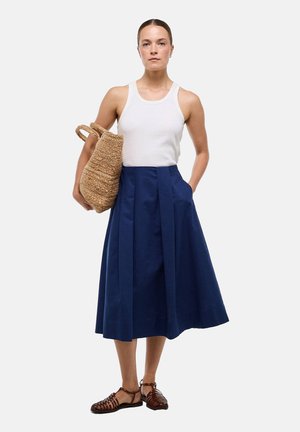 Femme portant un haut blanc sans manches, une jupe midi bleu marine, des sandales tressées marron, tenant un grand sac cabas tressé sur son bras.