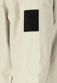 Beige Outerwear mit einem schwarzen rechteckigen Patch mit der Aufschrift "SLOPE STYLE", glatte Textur, minimale sichtbare Nähte und strukturiertes Design.