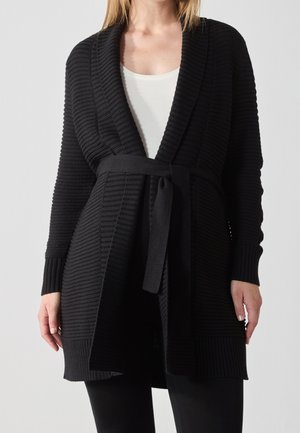 Kvinde iført en sort ribstrikket cardigan med sjalskrave og bundet taljebælte over en hvid top og sorte bukser.