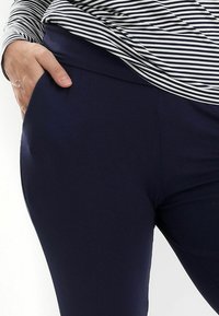 Marineblaue Leggings mit einer glatten, dehnbaren Textur und einem elastischen Bund. Eine Hand ist in eine seitliche Tasche in der Hüfthöhe gelegt.