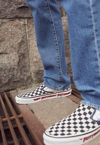 Baskets slip-on à carreaux noirs et blancs avec une semelle blanche et le texte rouge "FAST TIMES" sur le côté, associées à un jean en denim bleu.