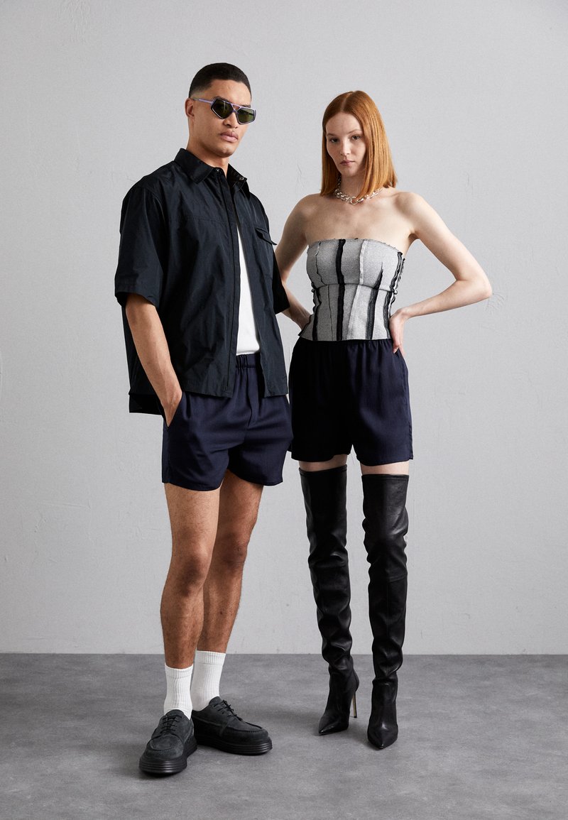 Schwarzes Übergrösse-Hemd, weisses T-Shirt, marineblaue Shorts und dunkle Sneakers bei einem Mann; Frau in einem trägerlosen gestreiften Oberteil und hohen schwarzen Stiefeln.