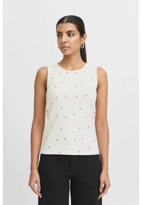 Top sans manches en côtelé beige clair avec des motifs de cœurs rouges éparpillés. Encolure ronde et silhouette ajustée. Associé à un pantalon noir.
