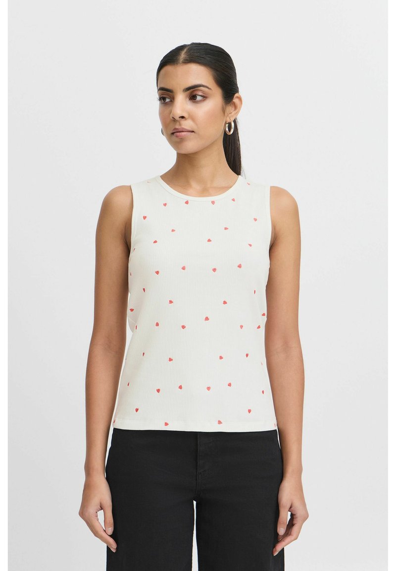 Top sans manches en côtelé beige clair avec des motifs de cœurs rouges éparpillés. Encolure ronde et silhouette ajustée. Associé à un pantalon noir.