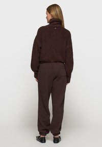 Brązowy, puszysty sweter z wysokim kołnierzem, w połączeniu z ciemnobrązowymi spodniami dresowymi z elastycznymi mankietami i czarnymi butami typu slip-on. Minimalistyczny design.