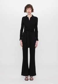 Combinaison noire à manches longues, bustier ajusté et jambes évasées. Dotée d'un col à revers boutonné et d'une ceinture en tissu à la taille. Texture lisse.