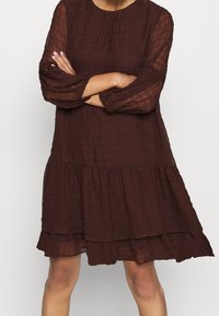 Kvinna med korsade armar som bär en lös, lager-på-lager, strukturerad långärmad klänning i mörkbrun färg, stående mot en enkel ljus bakgrund.