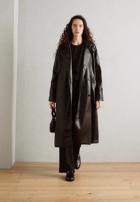 Abrigo negro largo en material suave, diseño cruzado, con un cinturón desmontable. Combinado con pantalones y zapatos negros.