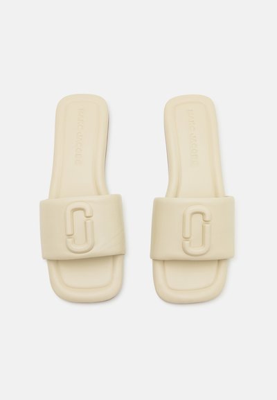 Beige slide sandals featuring a wide, textured strap, embossed logo detail, and flat sole design. Made of soft, smooth material.