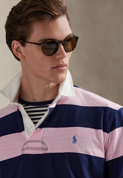 Tumesinise ja roosa triibulise kraega polo särk, millel on valge krae ja väike sinine tikitud logo. Mustad päikeseprillid kilpkonnakoorraamistikus.