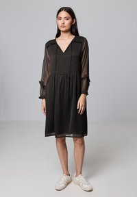 Vestido negro transparente con escote en V, mangas largas y acentos con volantes. Presenta una cintura fruncida y un patrón texturizado sutil. Se lleva con zapatillas blancas.