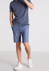 Homme portant un polo rayé bleu et blanc, un short en denim bleu clair et des baskets blanches, se tenant avec une main dans sa poche.